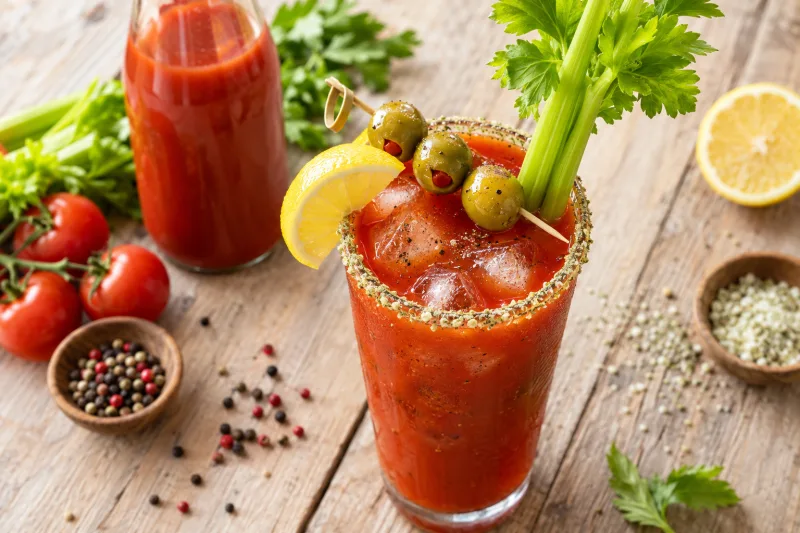 Bloody Mary Cocktail mit Staudensellerie, Zitrone und Oliven, auf Holztisch, Eiswürfel sichtbar.