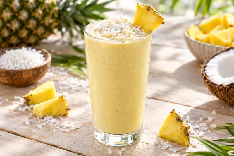 Veganer Kokos-Ananas-Smoothie im Glas, garniert mit Ananasstück und Kokosraspeln, auf hellem Hintergrund.