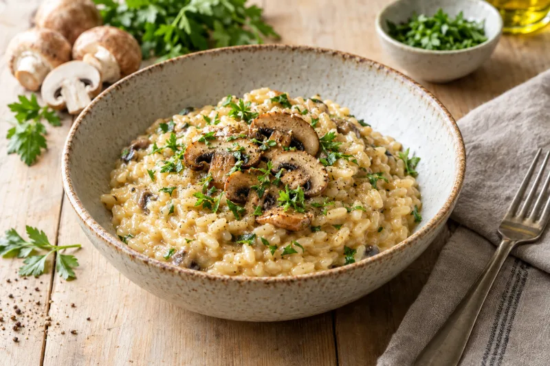 Veganes Pilz-Risotto in einer weißen Schüssel, garniert mit frischer Petersilie und einigen Pilzscheiben, daneben ein Löffel.
