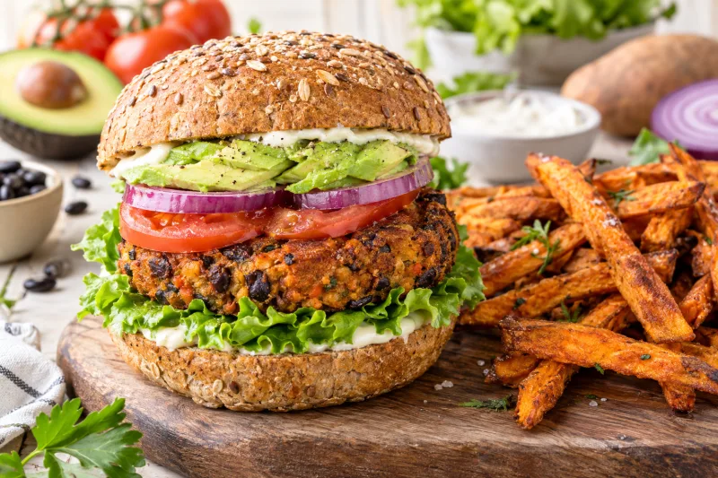 Veganer Süßkartoffel-Burger mit schwarzen Bohnen, Avocado, Tomate und Salat auf einem Holzbrett