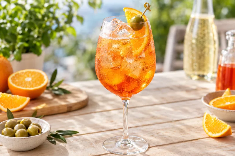 Veganer Aperol Spritz in großem Weinglas mit Eis, Orangenscheibe und grüner Olive