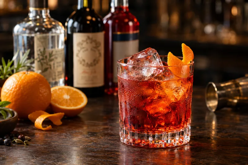 Veganer Negroni Cocktail in einem Tumbler mit Eiswürfeln und Orangenzeste, auf einer Bar mit Gin, Wermut und Campari im Hintergrund.