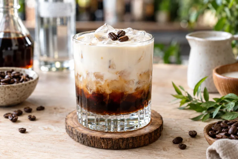 Veganer White Russian Cocktail in einem Tumbler mit Eiswürfeln, cremiger Schicht und Kaffeebohnen auf Holzuntersetzer.