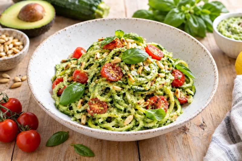 Zucchini-Nudeln mit cremigem Avocado-Pesto, garniert mit Kirschtomaten und Pinienkernen auf weißem Teller