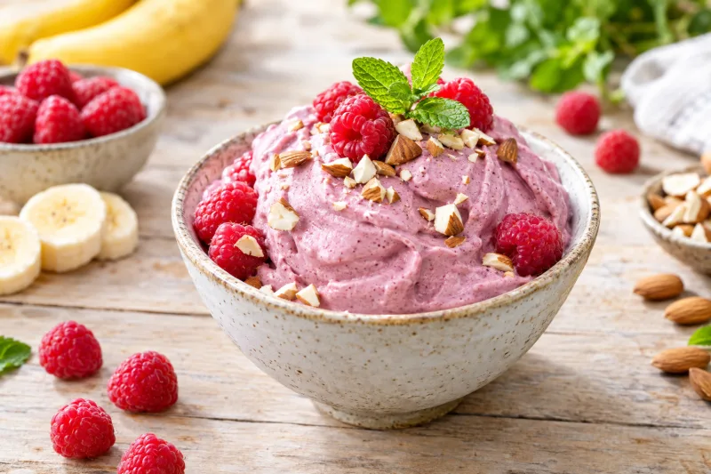 Cremige lila Bananen-Himbeer-Nicecream in einer Schale mit frischen Himbeeren und Mandeln garniert