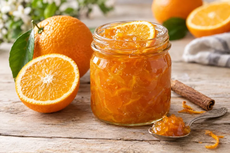 Glas mit veganer Orangenmarmelade, daneben frische Orangen und ein Löffel