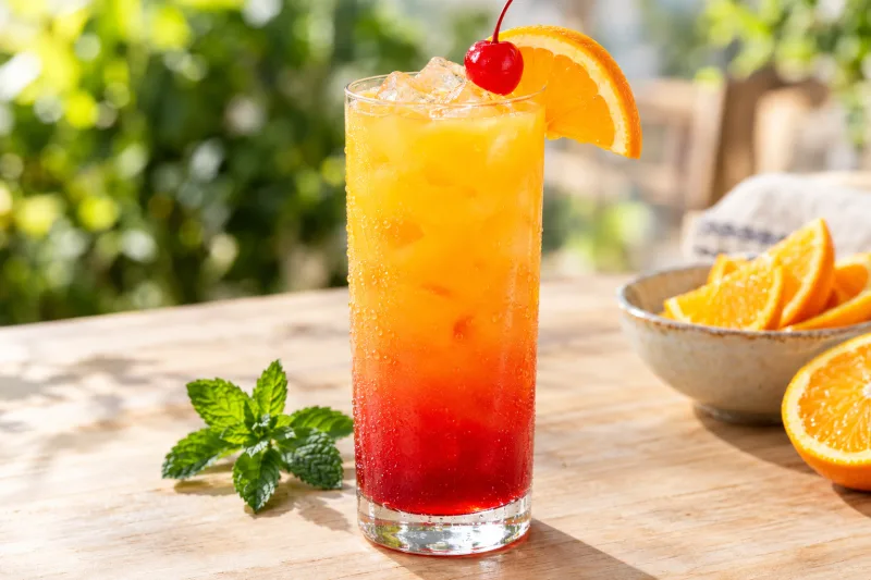 Veganer Tequila Sunrise Cocktail im hohen Glas mit Farbverlauf, Orangenscheibe und Kirsche